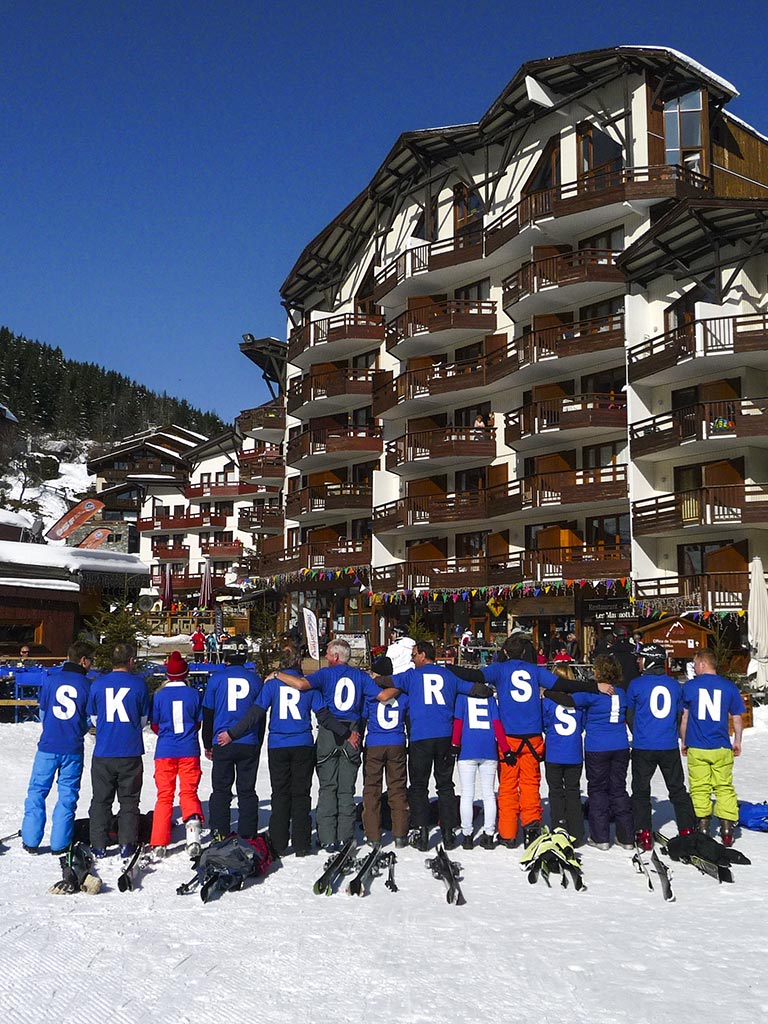 group of people seen from behind, each with a letter written on their t-shirt, all forming the words: ski progression